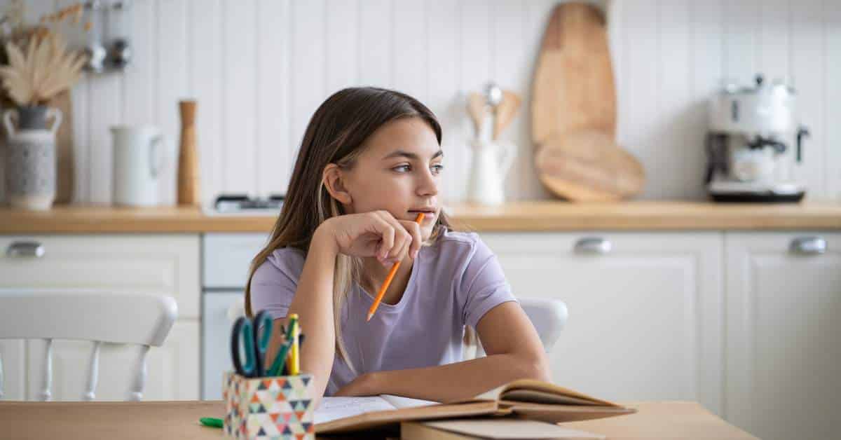 Teen girl looking out the windows. She has a pencil in her hand and a notebook in front of her