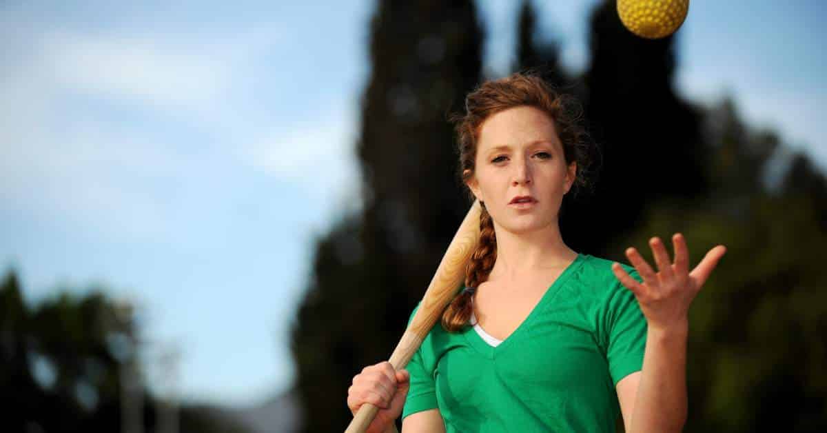 Teen girl wearing a green jersey and holding a softball bat and softball.