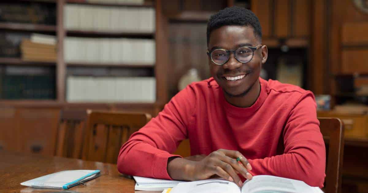 College student sitting at a desk with his books in front of him. He is looking at the camera and smiling.