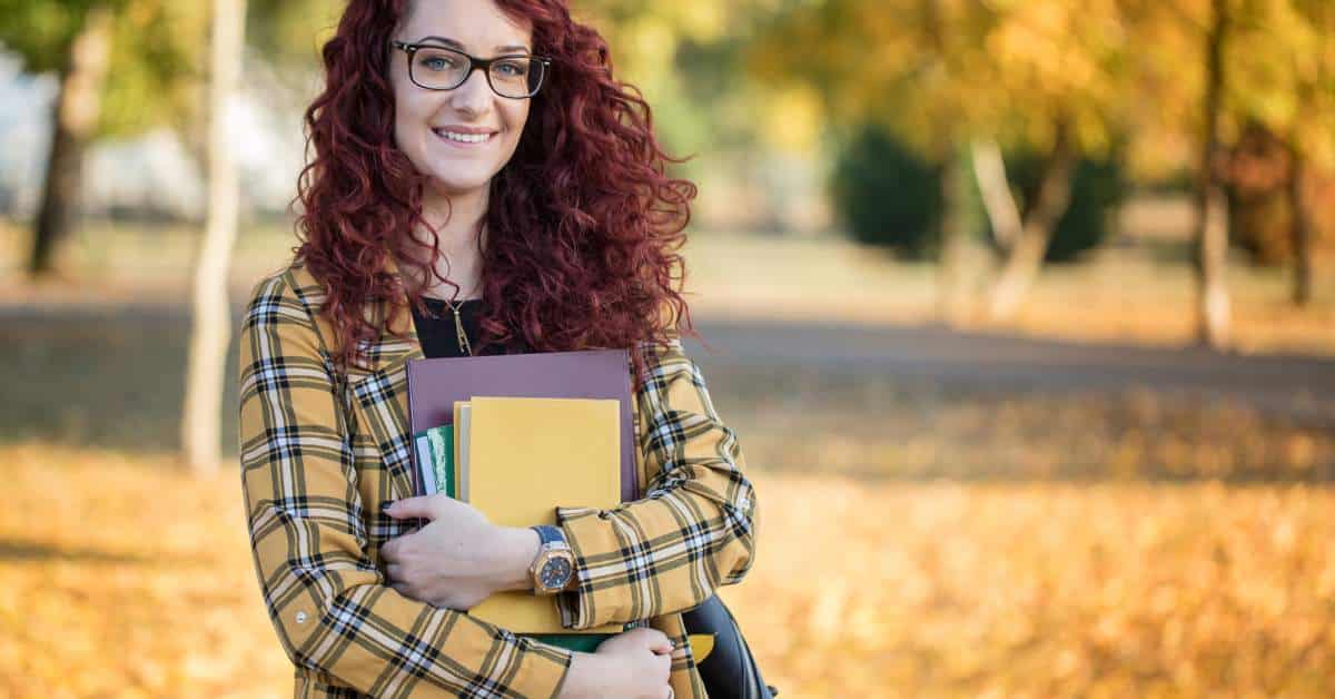 College student with curly red hair and glasses holding notebooks to her chest and smiling at the camera.