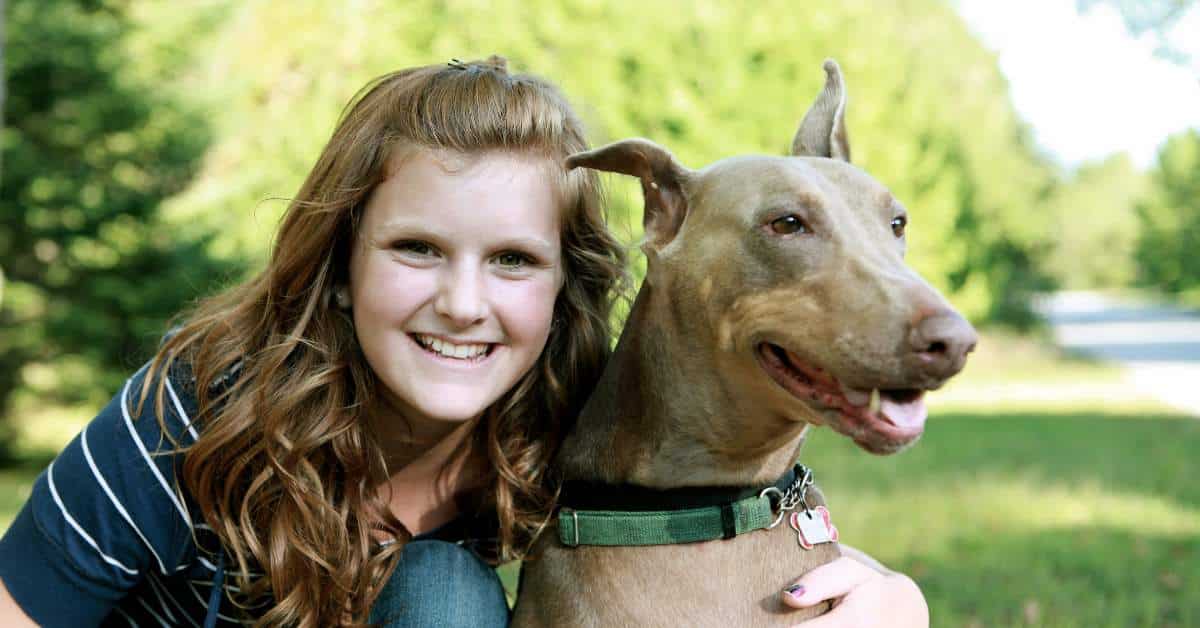 Teen girl with her arm wrapped around her dog. She's smiling into the camera, and the dog is looking off in the distance.