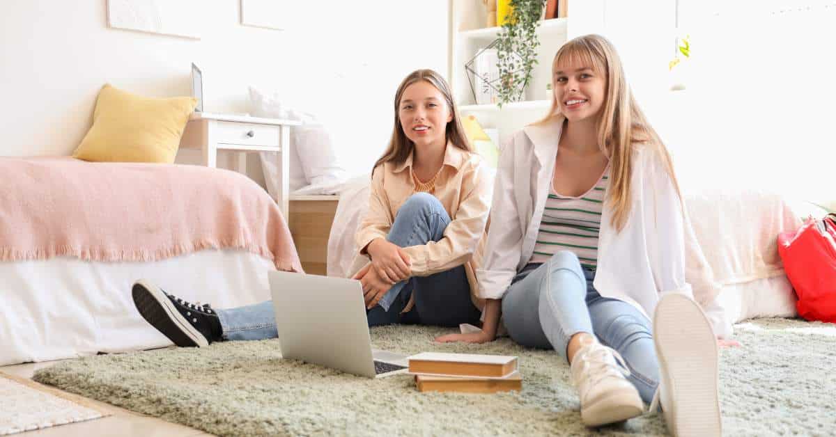 Two girls in a pink and white dorm room sitting on a carpet, smiling at the camera.