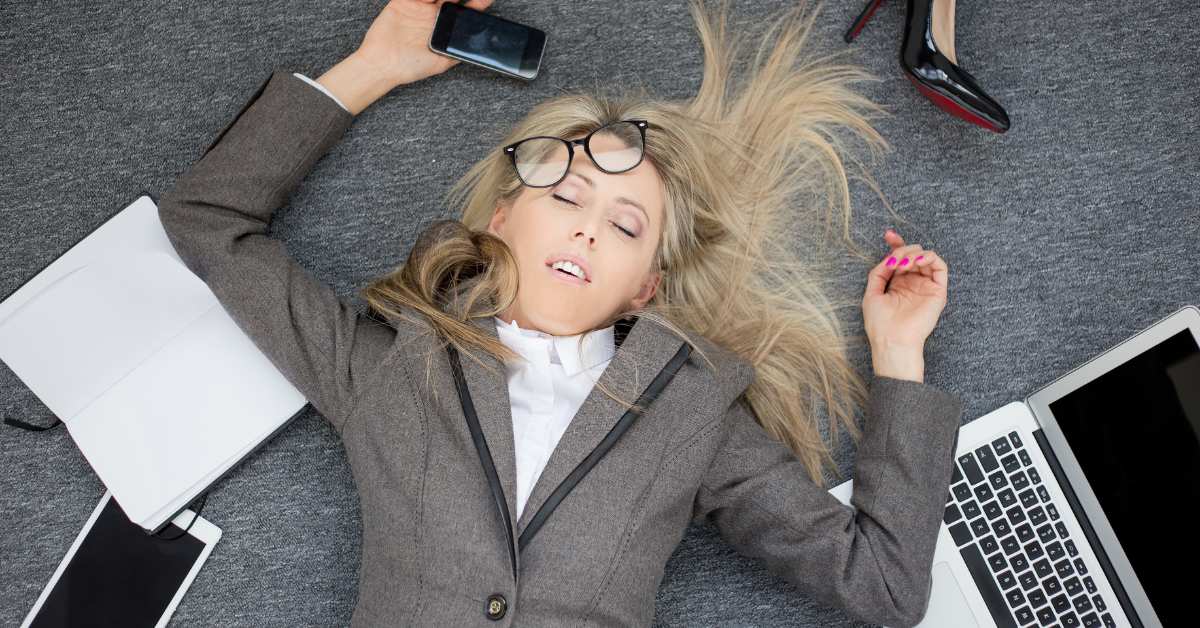 Exhausted woman lying on the floor with her eyes closed, holding a cell phone with a notebook and laptop by her