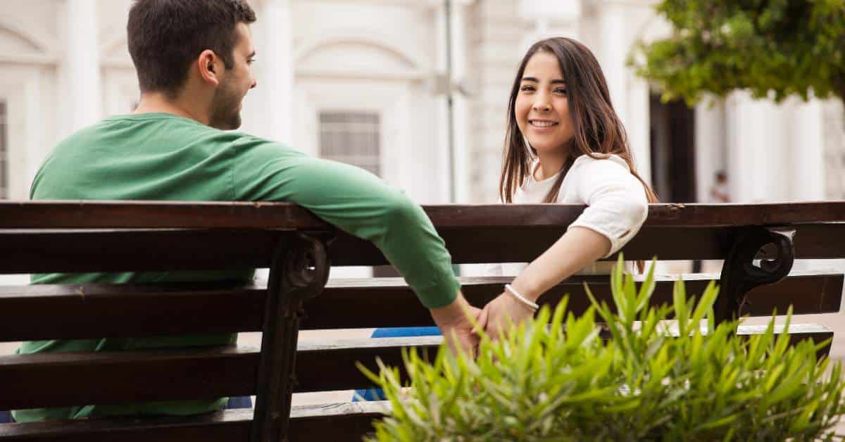 Teen guy and girl sitting on a park bench holding hands. He's looking at her; she's looking back at the camera.