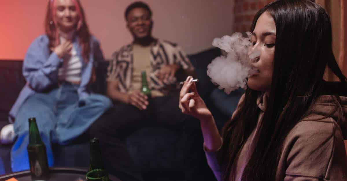 Teenage girl smoking marijuana while another teen girl and a teen boy look on in the background.