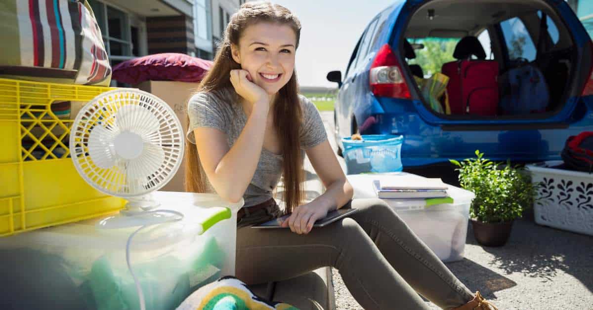Female sitting near boxes and a fan while her parents unpack the car. They are moving her into her dorm room.