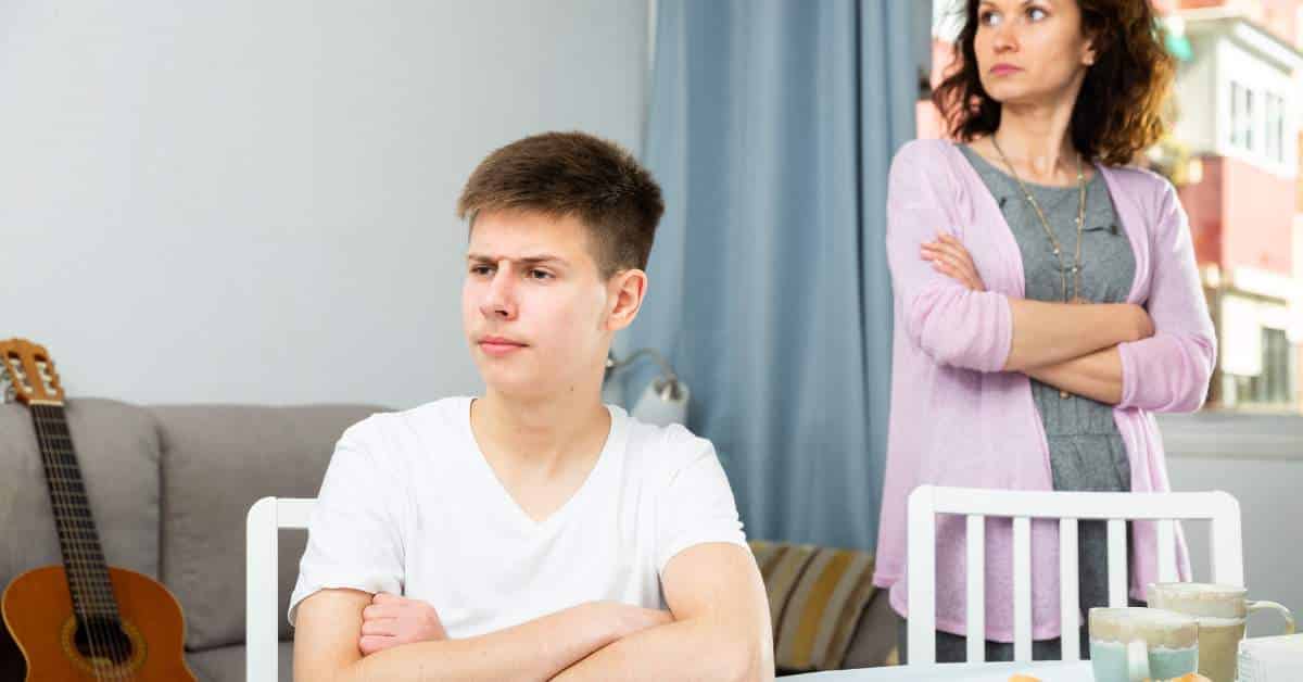 Mother in the background with her arms crossed while teen son is in the foreground, back to mom, with his arms crossed across his chest.