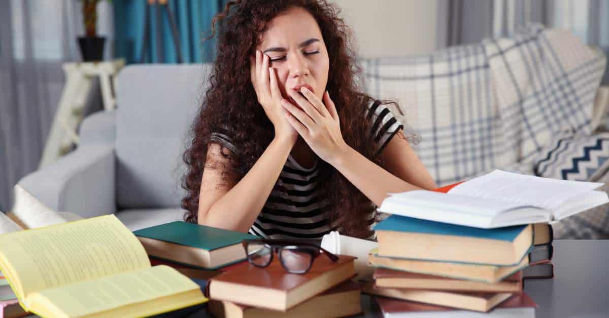 Exhausted teen girl stifles a yawn. She is surrounded by piles of school books.