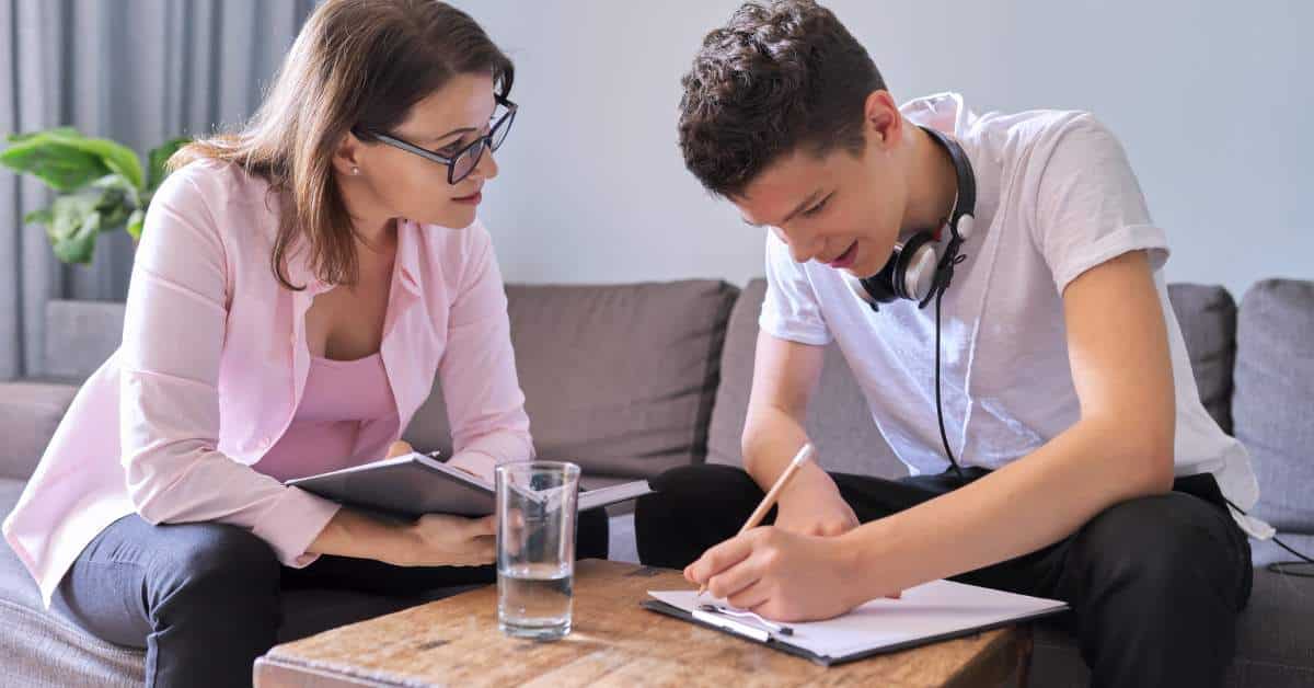 Teen boy writing on a clipboard on top of a table. His mother leans in, smiling at him, and holds a book.