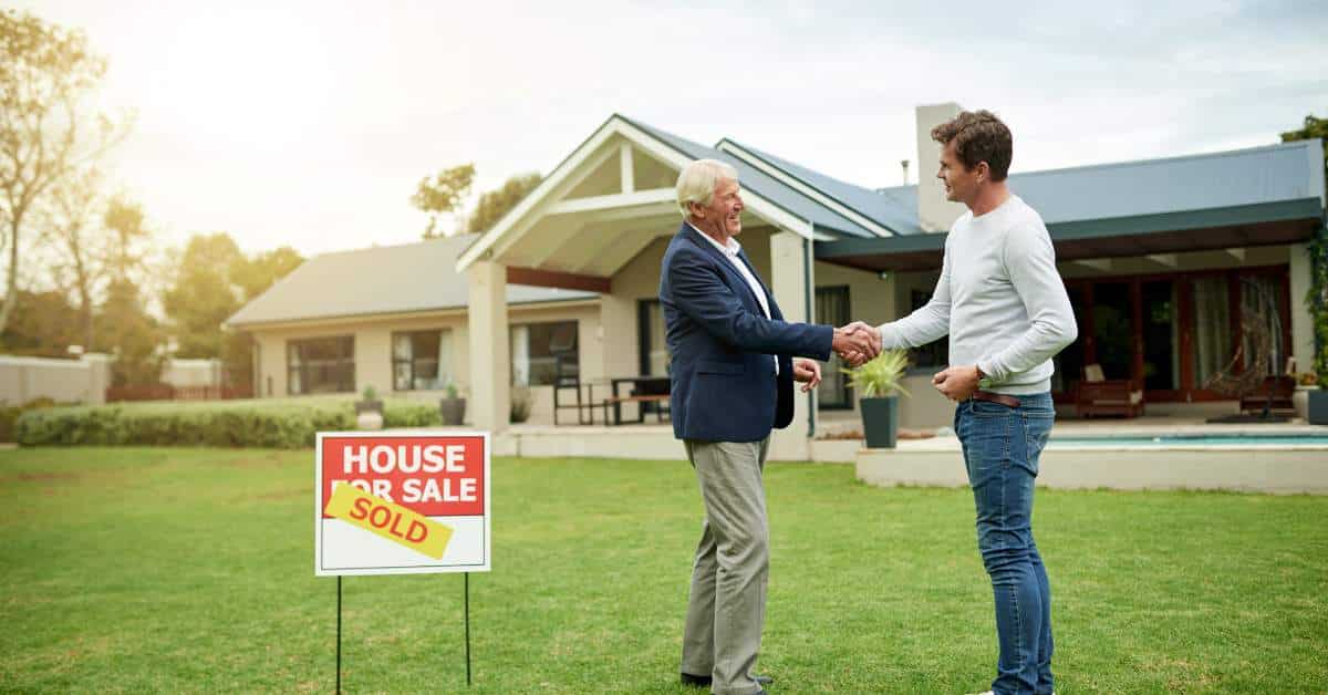 Young man shaking hands with a realtor with white hair. There is a house in the background and a for sale sign covered with a "sold" sign.