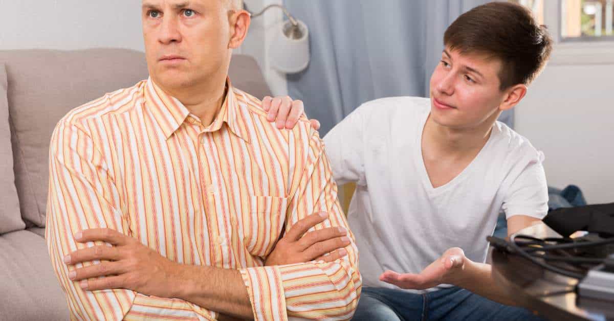 Father is in the forefront with his arms crossed across his chest. His back is to his teen son who has one hand on his shoulder and the other hand outstretched.