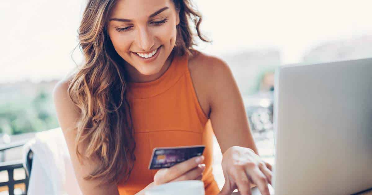 Young woman with long hair has a smile on her face as she looks down at her credit card and has a laptop open in front of her