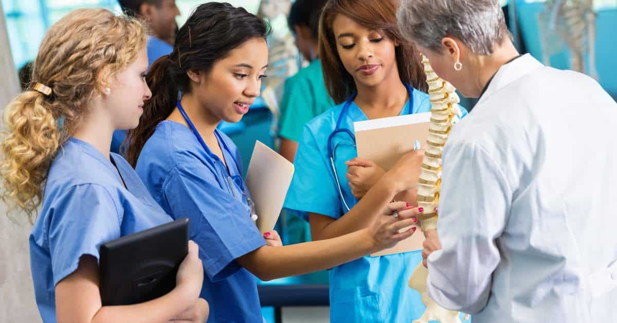 Three female nursing students in scrubs examing a skeleton's spine while their teacher, in a white coat, looks on.