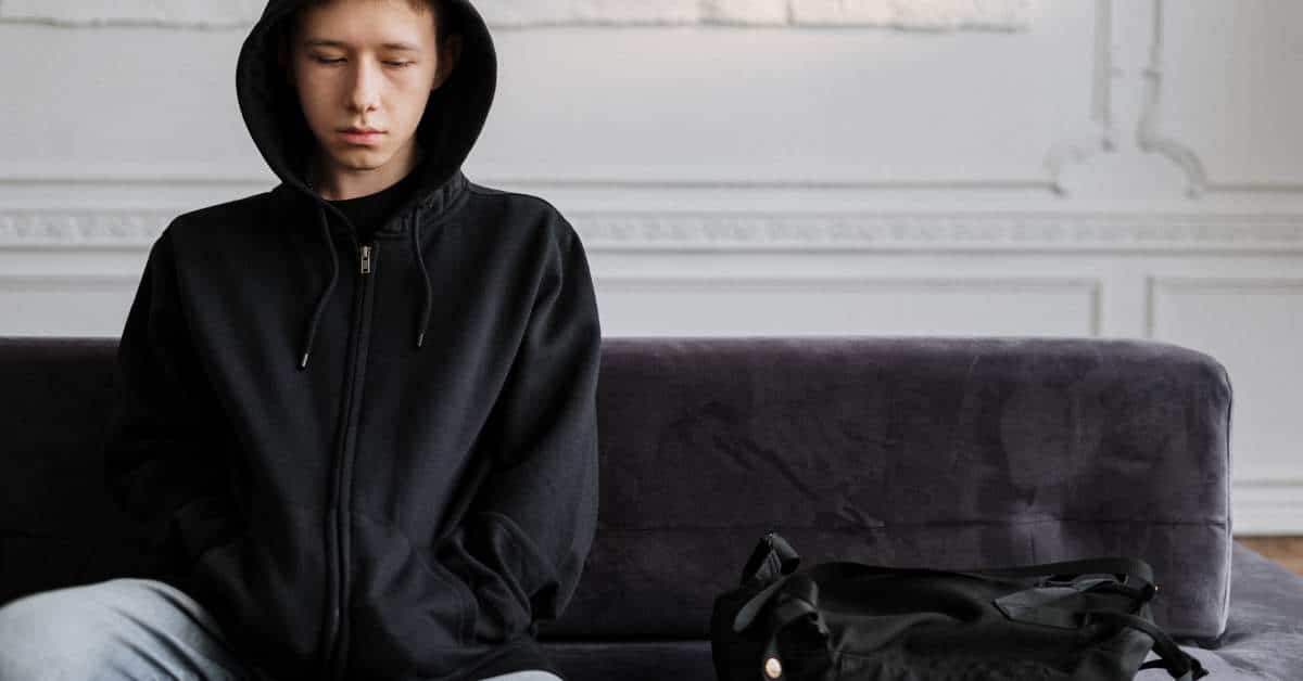 Teenage boy wearing a hoodlie sitting on a couch with a bag beside him. He looks depressed.