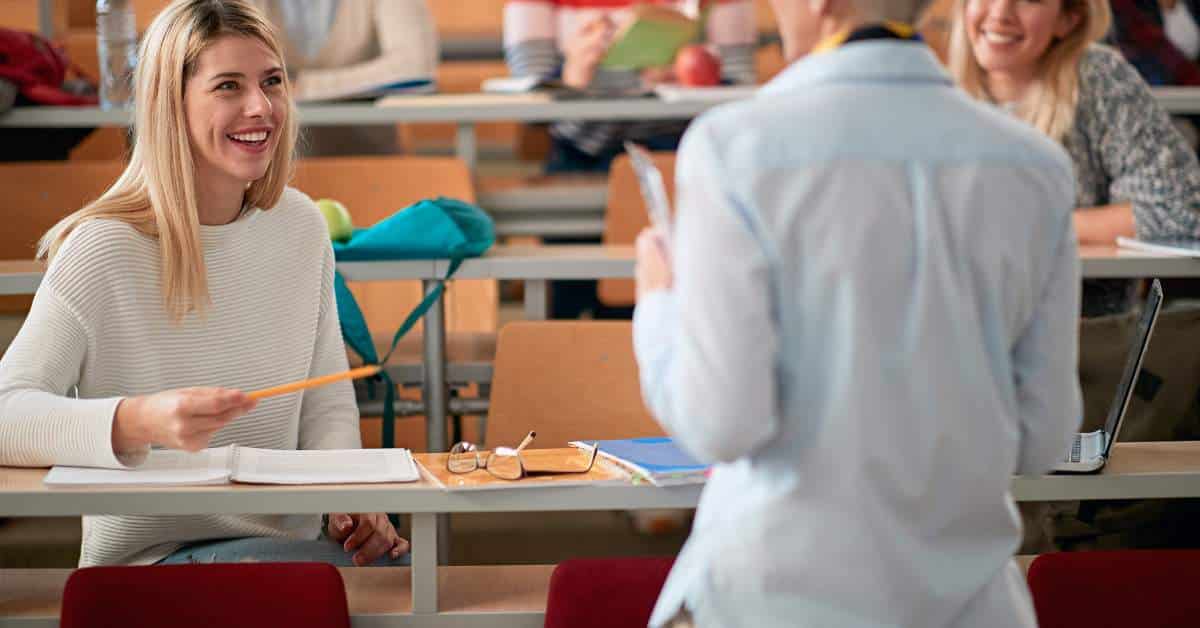 Young woman sitting at a desk smiling up at professor