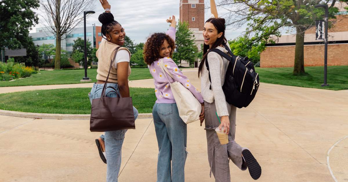 Three female college students turn back to the camera smiling and lifting their hands in the air.