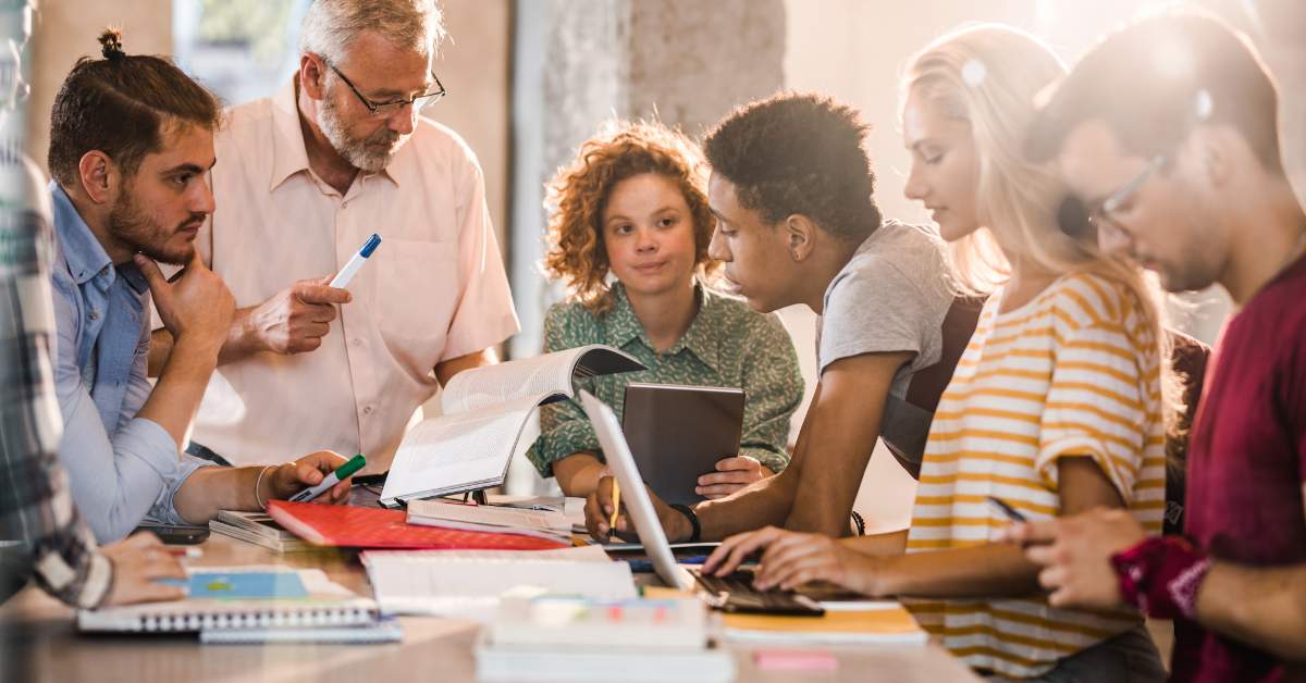 A college professor with gray hair consults with students seated around a desk