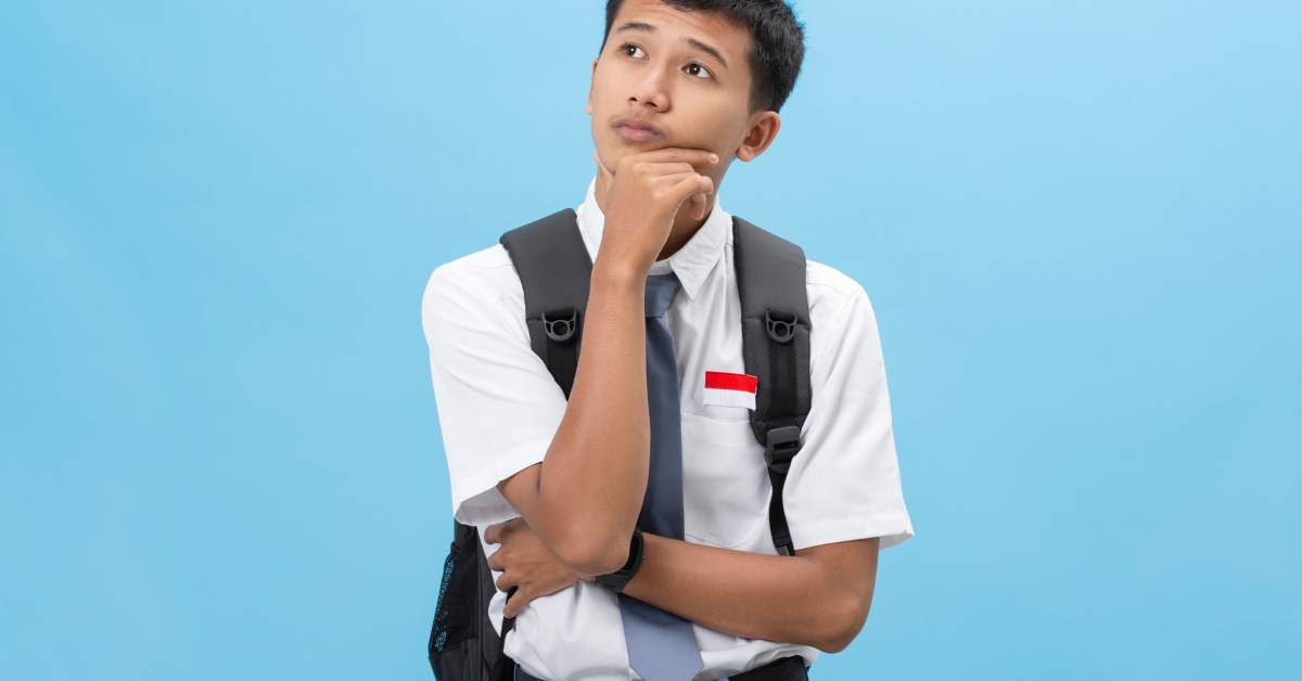 Young Asian man wearing a backpack and a white shirt and tie, holds his chin and looks up, confused.