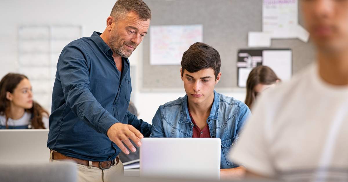 A middle aged professor stands and helps a young college student who is working at a computer.