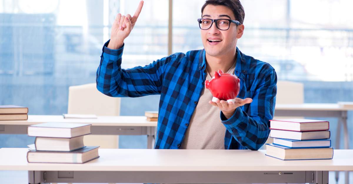 Man in a plaid shirt sitting at a desk holding a piggy bank in one hand and point up with his other hand.