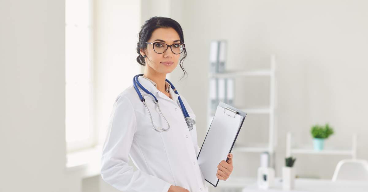 Woman wearing glasses, holding a clipboard, and wearing a white lab coat.
