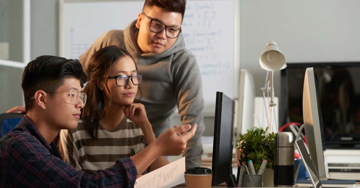 Three Asian students, two male and one female, huddling together looking at a computer screen.